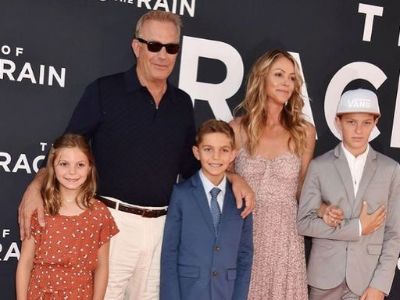 Grace Avery Costner is wearing a red dress, Cayden and Hayes Costner is in a suit, Christine Baumgartner is wearing a peach dress and Kevin Costner is wearing a black shirt and white pants.